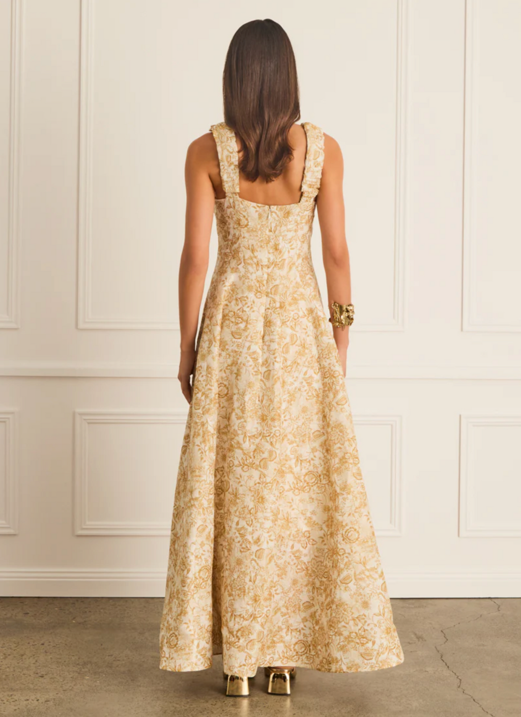 Woman wearing a long floral dress against a white paneled wall.