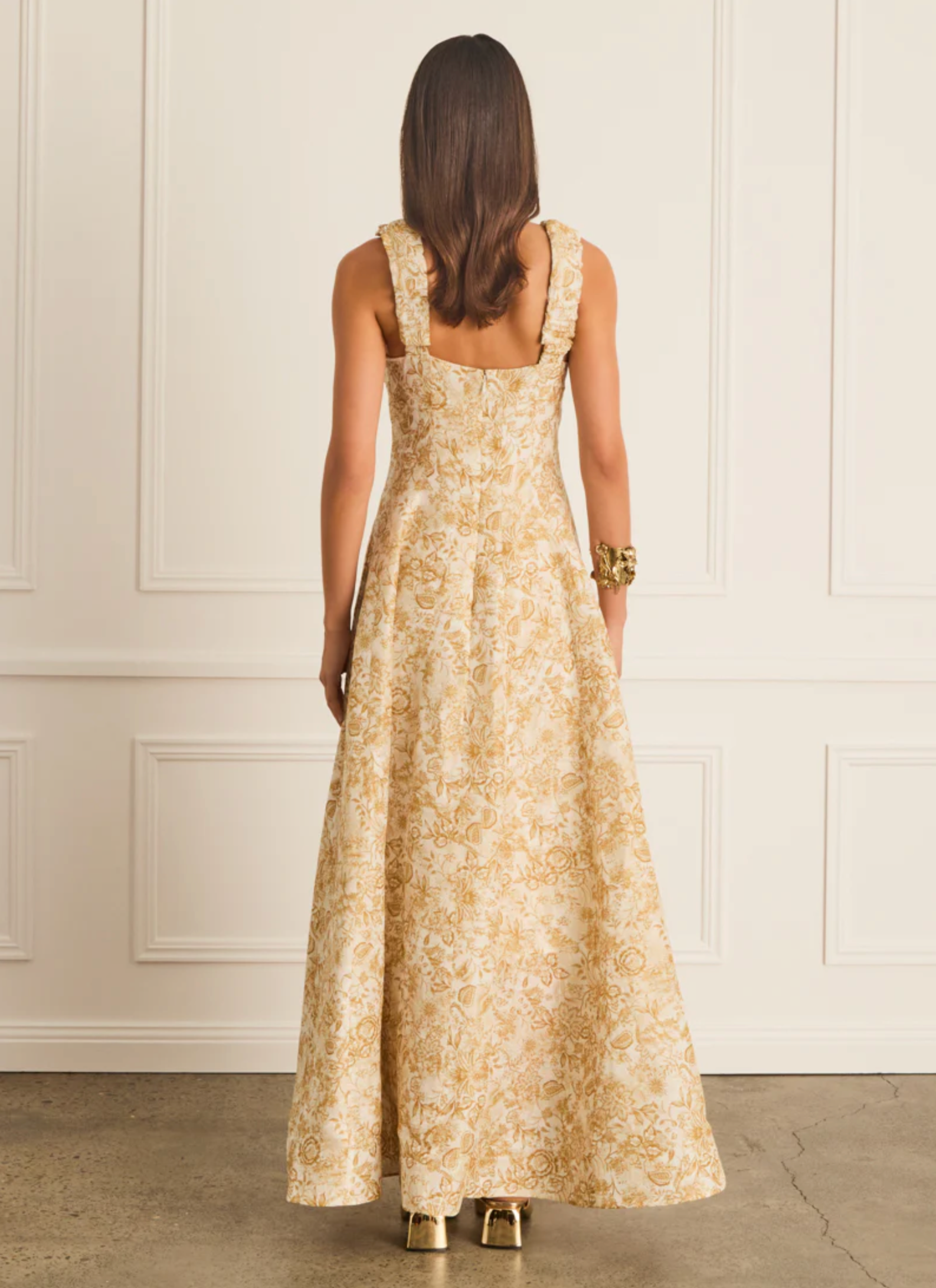 Woman wearing a long floral dress against a white paneled wall.