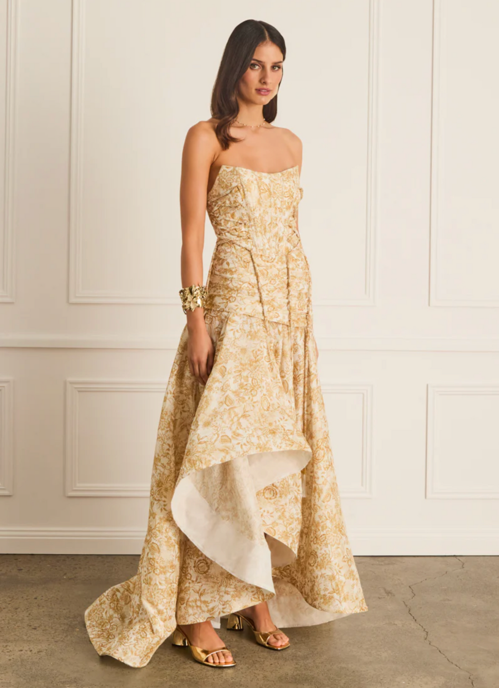 Woman wearing a gold strapless gown with a white underlayer against a white paneled wall.