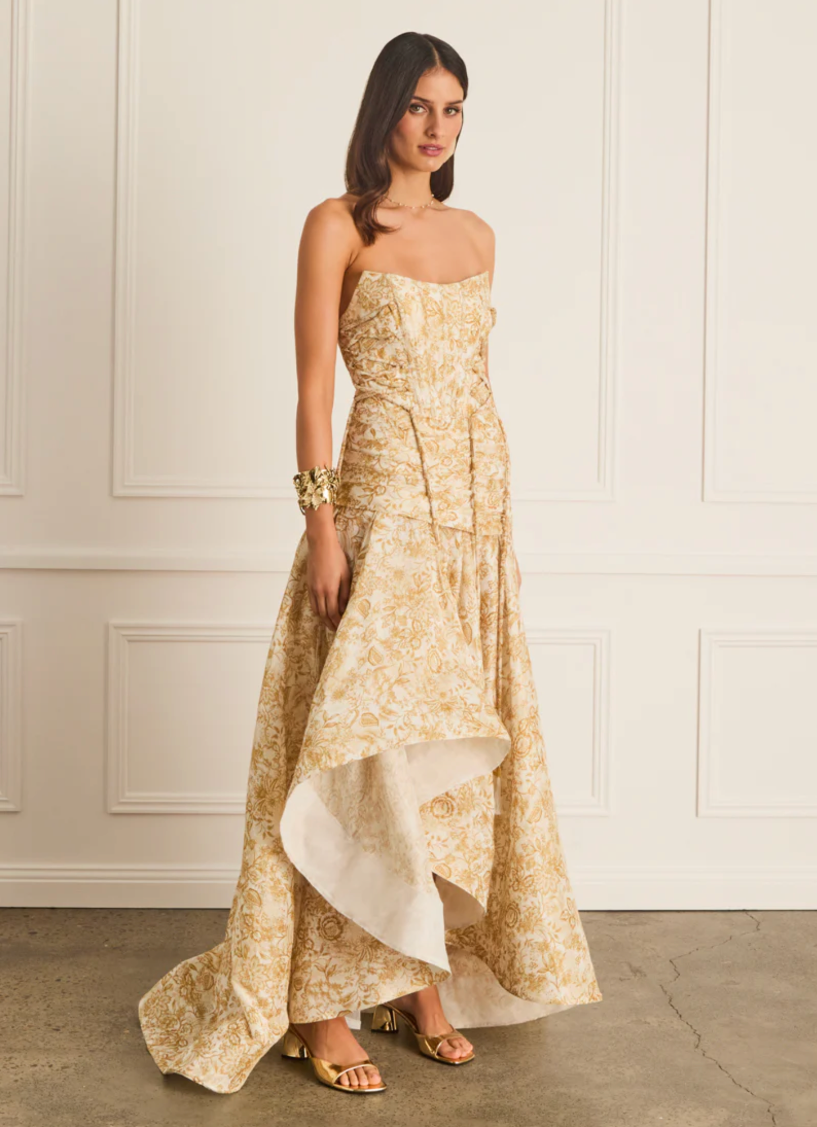 Woman wearing a gold strapless gown with a white underlayer against a white paneled wall.
