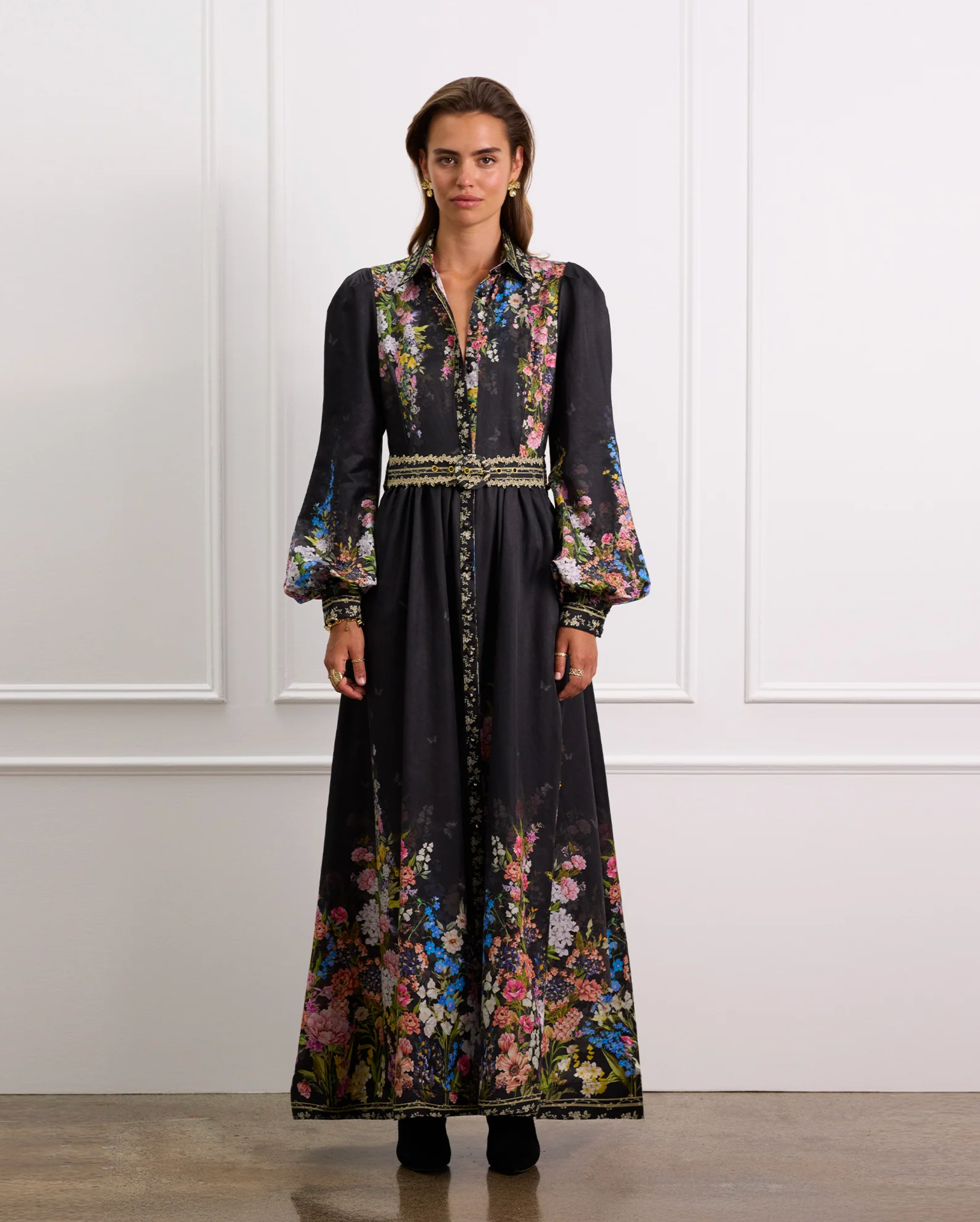Woman wearing a long, dark dress with floral patterns against a white wall.