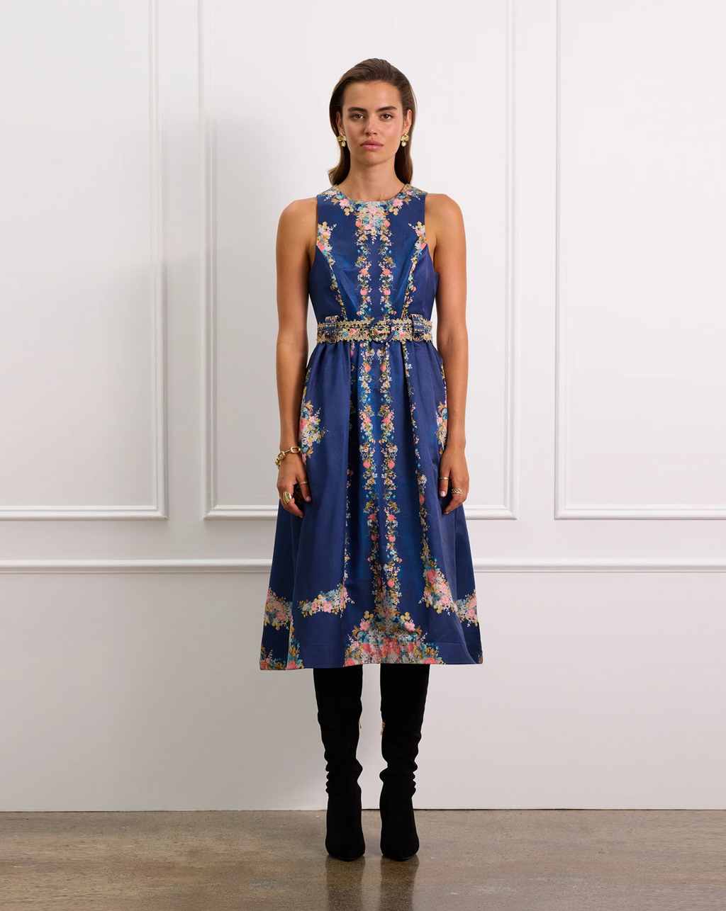 Woman wearing a blue floral dress with a belt against a white paneled wall.
