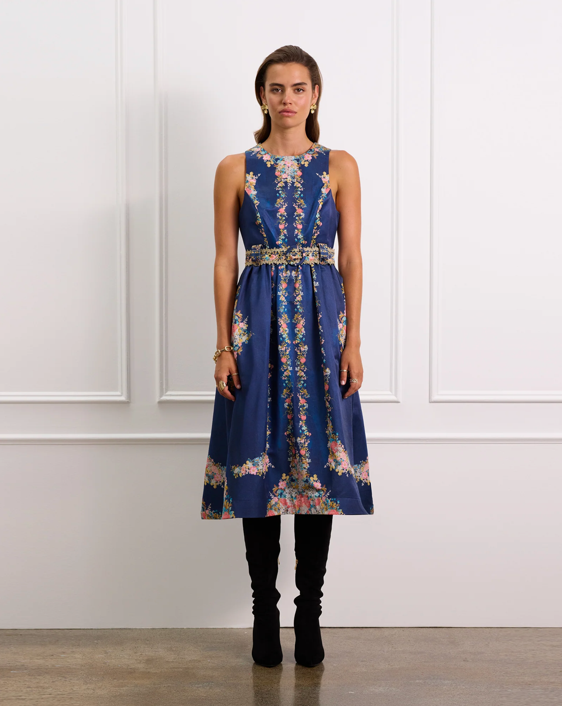 Woman wearing a blue floral dress with a belt against a white paneled wall.