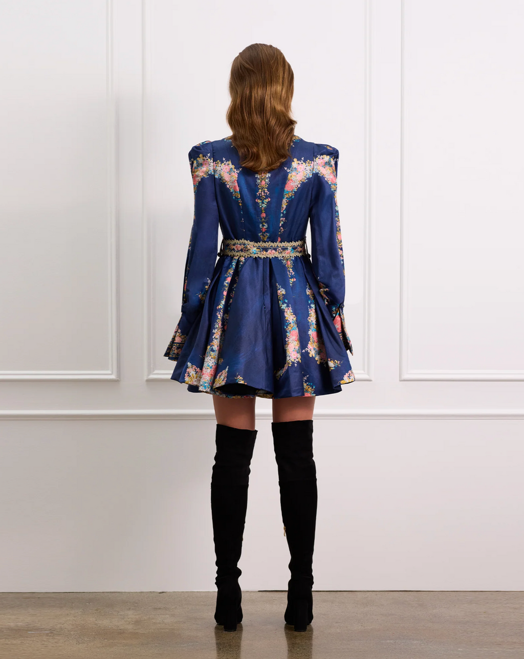 Woman wearing a blue floral dress with long sleeves and black knee-high boots against a white paneled wall.