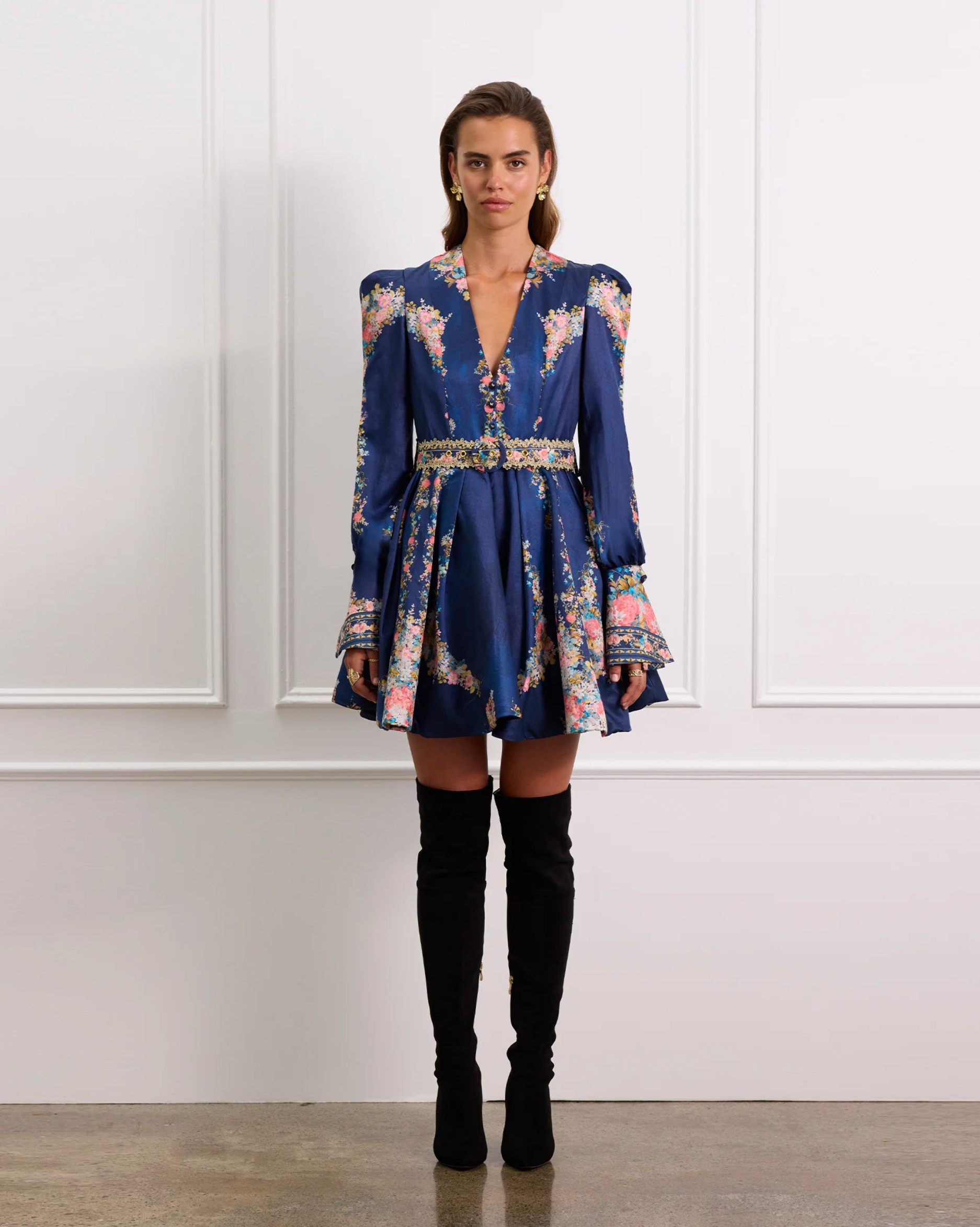 Woman wearing a blue floral dress with black knee-high boots against a white paneled wall.