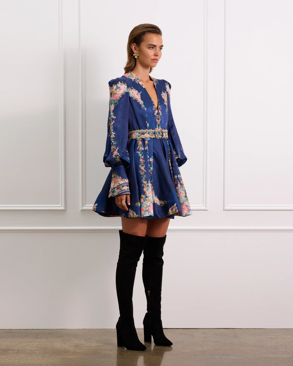 Woman wearing a blue floral dress with long sleeves and black knee-high boots against a white paneled wall.