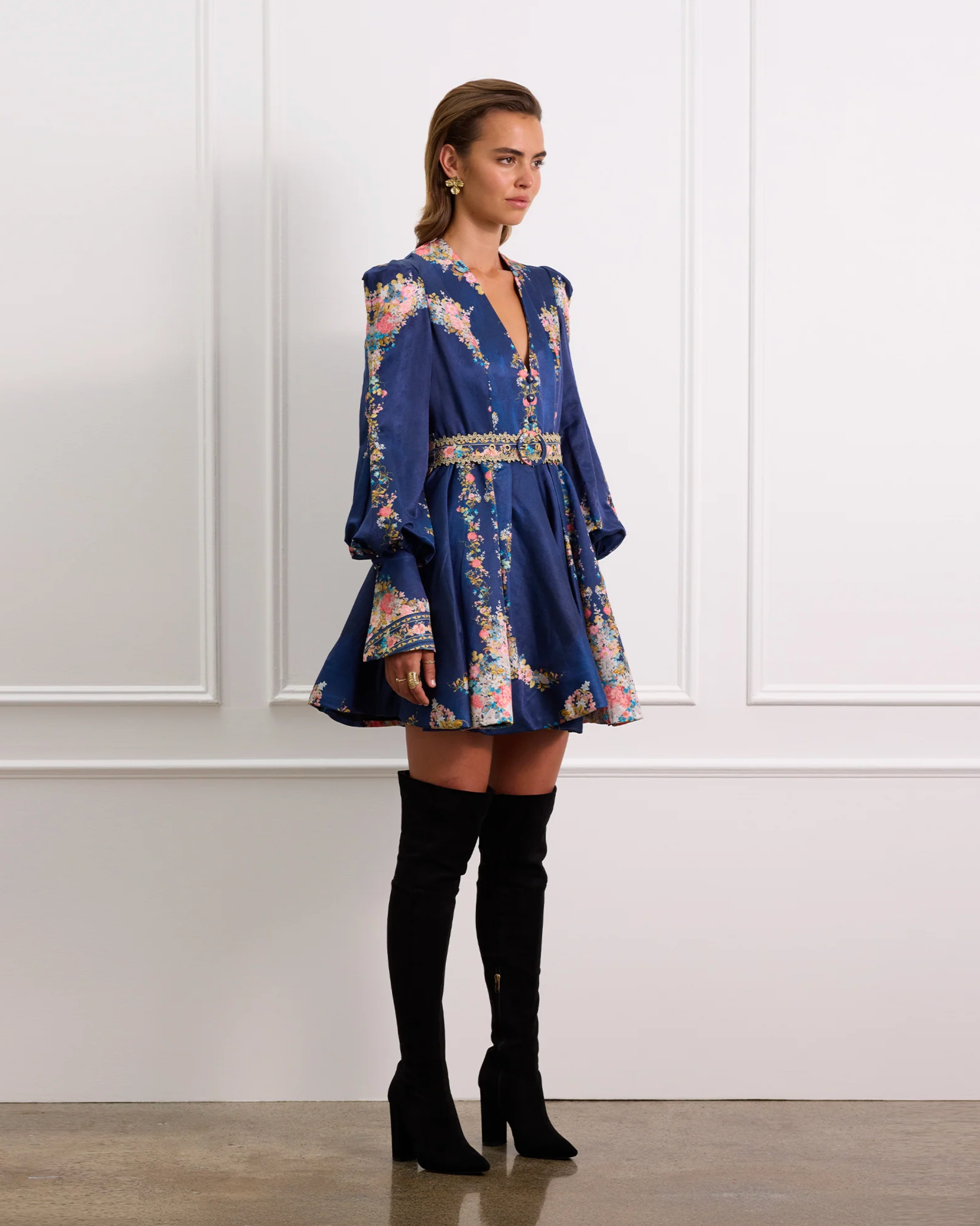 Woman wearing a blue floral dress with long sleeves and black knee-high boots against a white paneled wall.